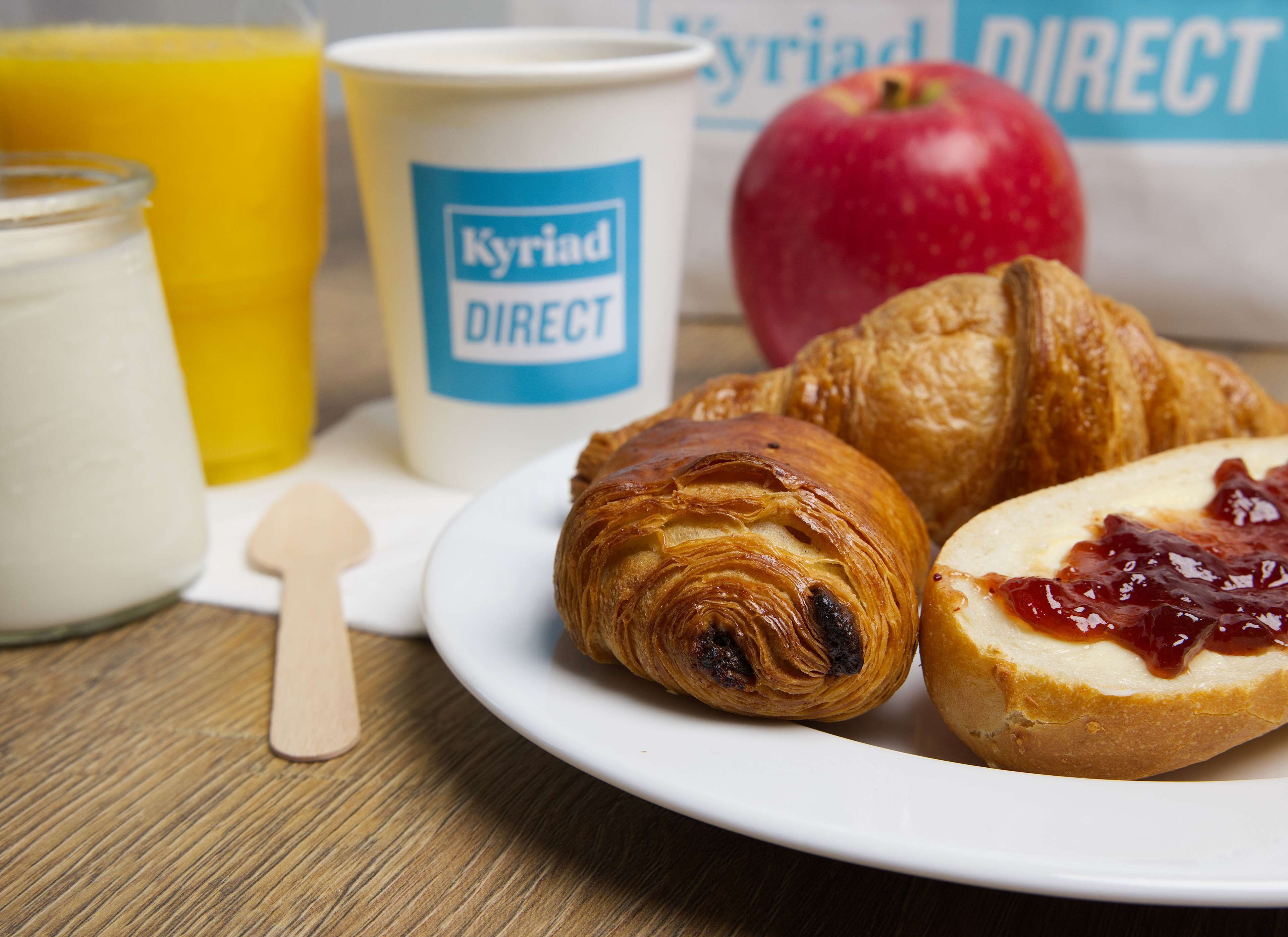 Ein Frühstücksteller mit Croissants und Marmelade, ein Glas Orangensaft, eine Milchkaraffe und eine Tasse mit dem Aufdruck 'Kyriad DIRECT'.