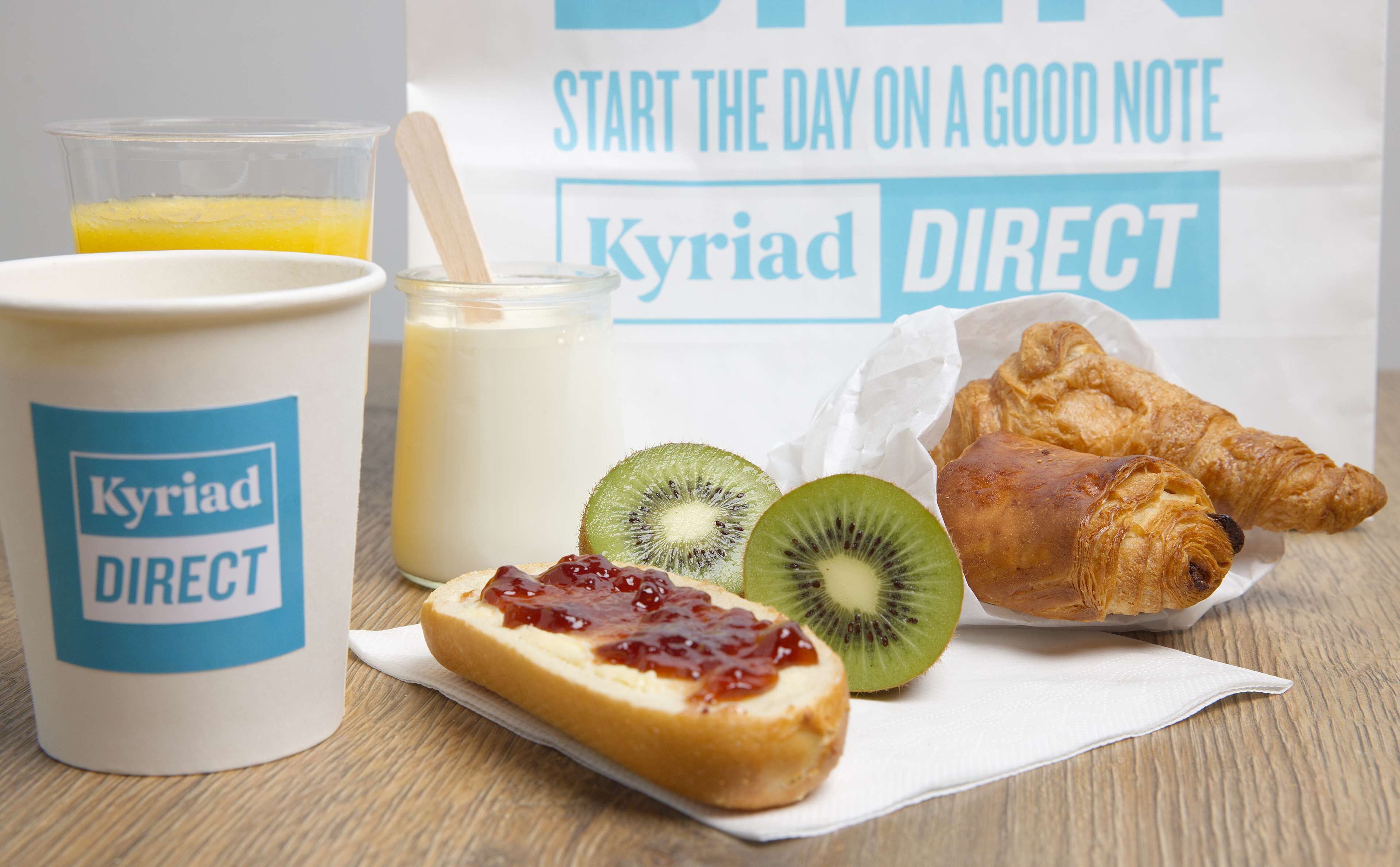 A breakfast setting with a cup labeled 'Kyriad DIRECT', a glass of orange juice, a jar of milk, a croissant, a slice of bread with jam, and a kiwi.