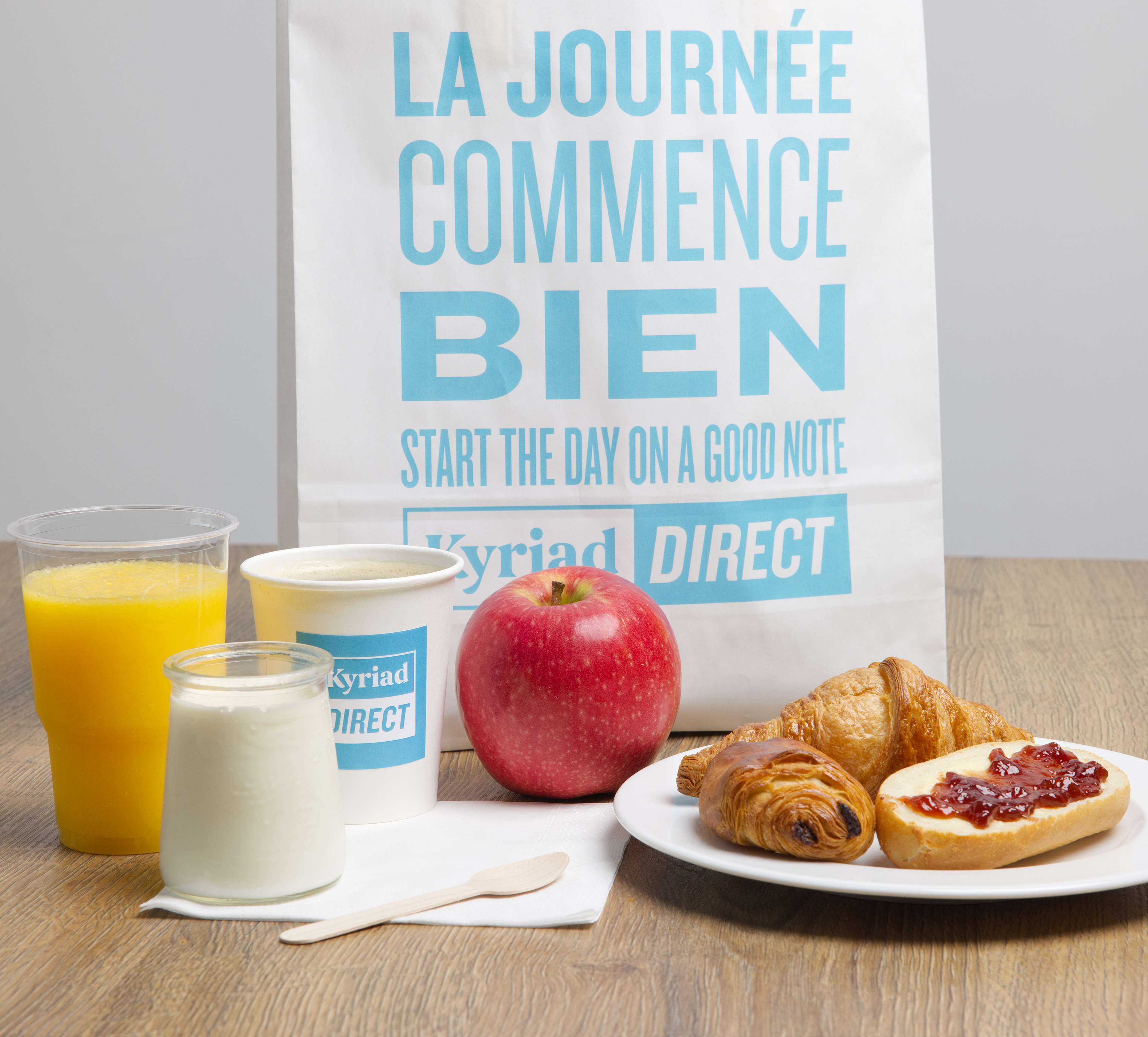 A breakfast setup with a bag, apple, croissants, and drinks on a wooden table.