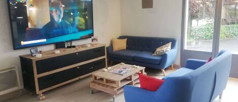 Un salón con un televisor de pantalla plana grande, un sofá azul, una mesa de centro de madera y una ventana con vistas a zonas verdes.