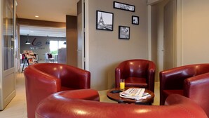 Un hall de hotel con sillones de cuero rojo, una mesa con un periódico y un vaso de jugo de naranja, y cuadros enmarcados en la pared.