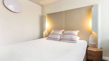 A neatly made bed with patterned pillows, a bedside lamp, and a wall-mounted mirror.