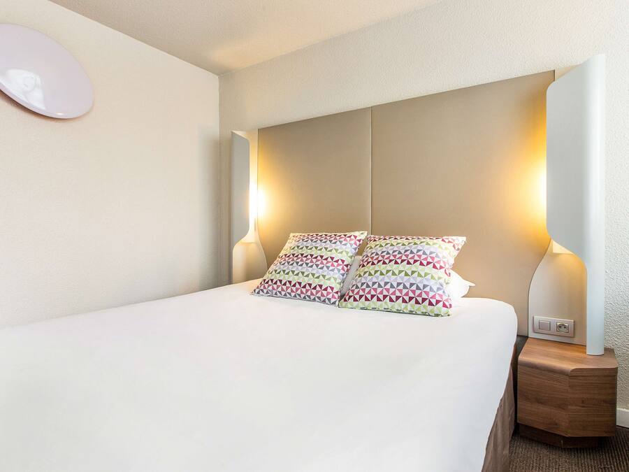 A neatly made bed with patterned pillows, a bedside lamp, and a wall-mounted mirror.