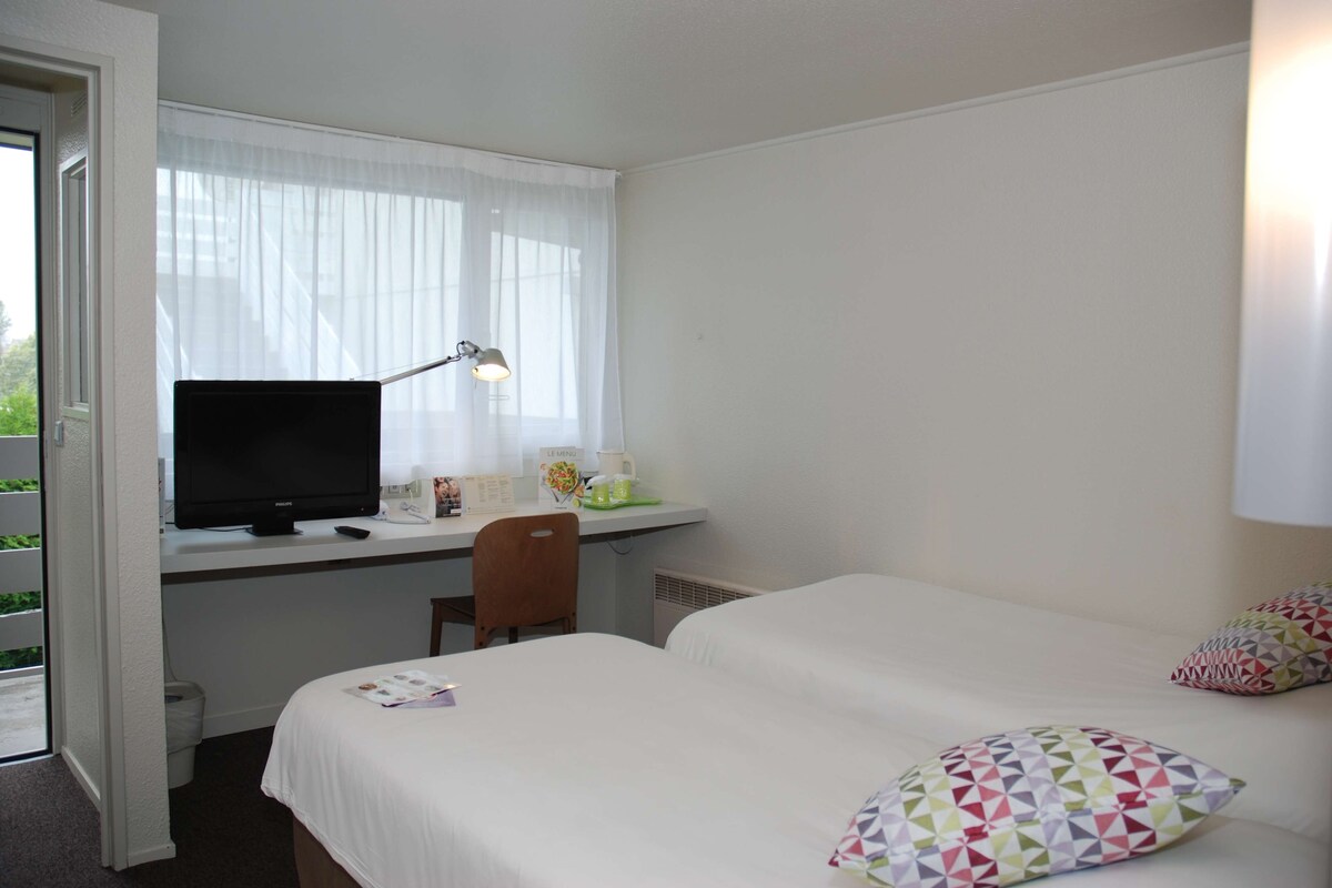 A hotel room with a bed, a desk with a computer and lamp, and a window with curtains.