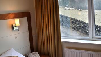 A hotel room with a bed, a bedside table with a lamp, and a view of a field with sheep.
