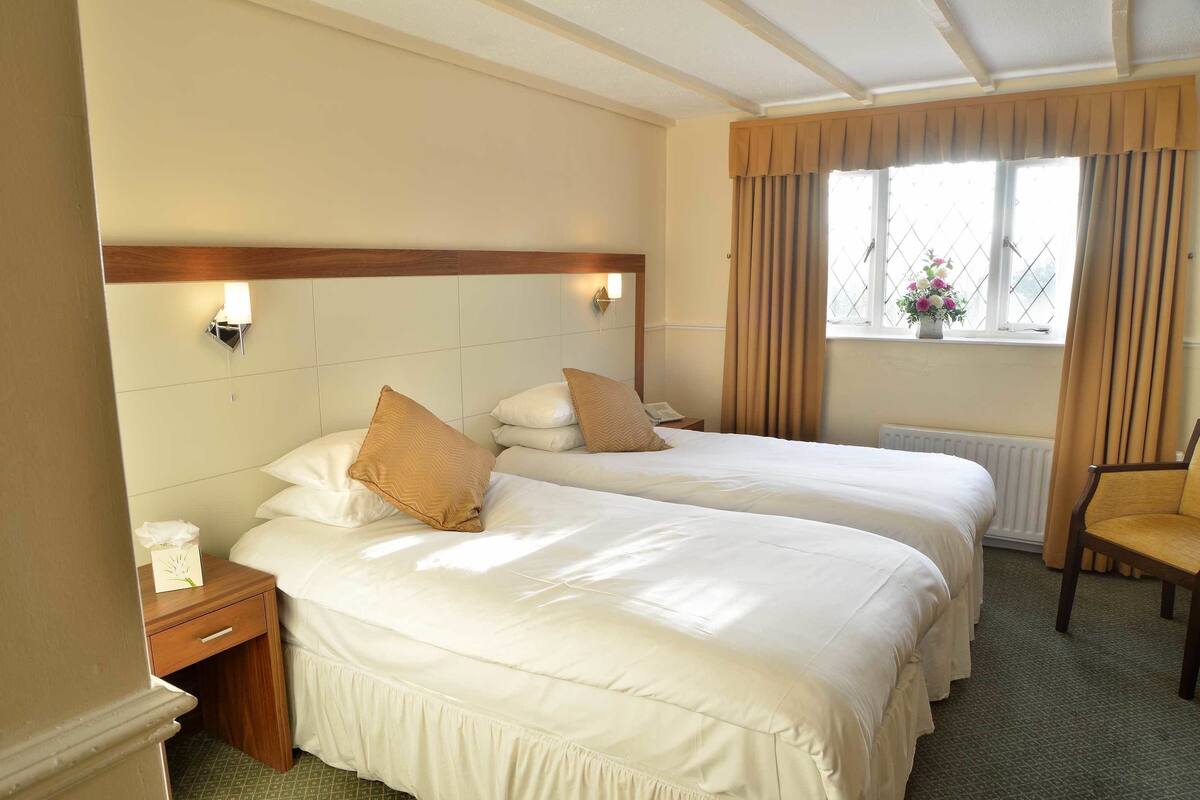 A hotel room with two beds, a wooden headboard, bedside lamps, a window with curtains, and a chair.