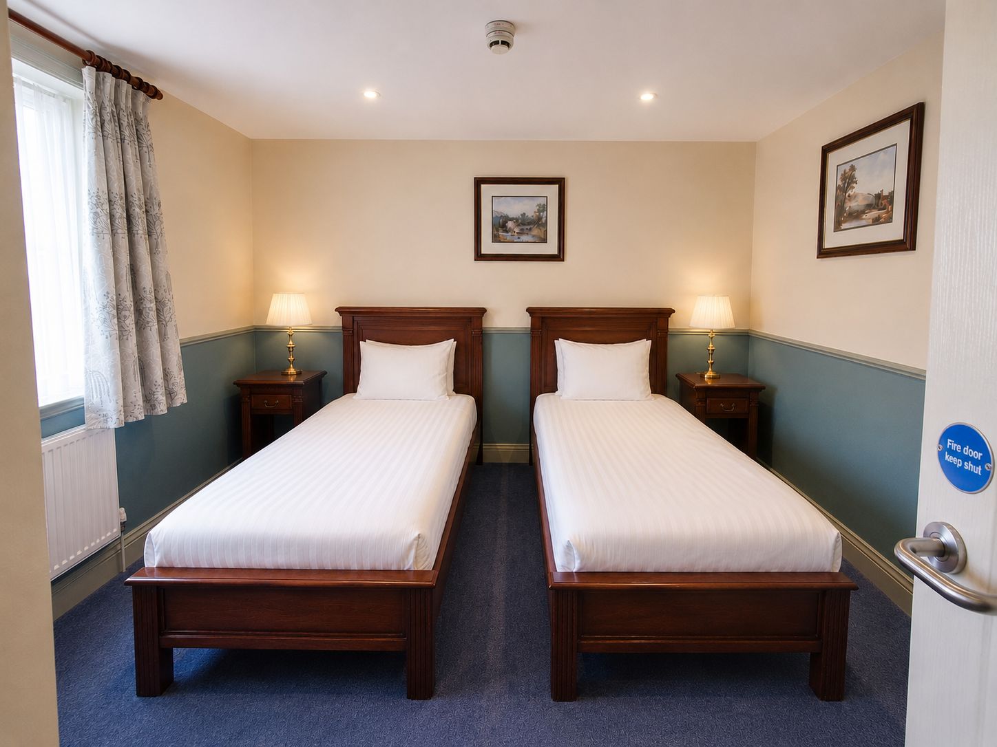 Habitación de hotel con dos camas individuales, mesitas de noche con lámparas, una ventana con cortinas y una puerta con letrero de seguridad contra incendios.