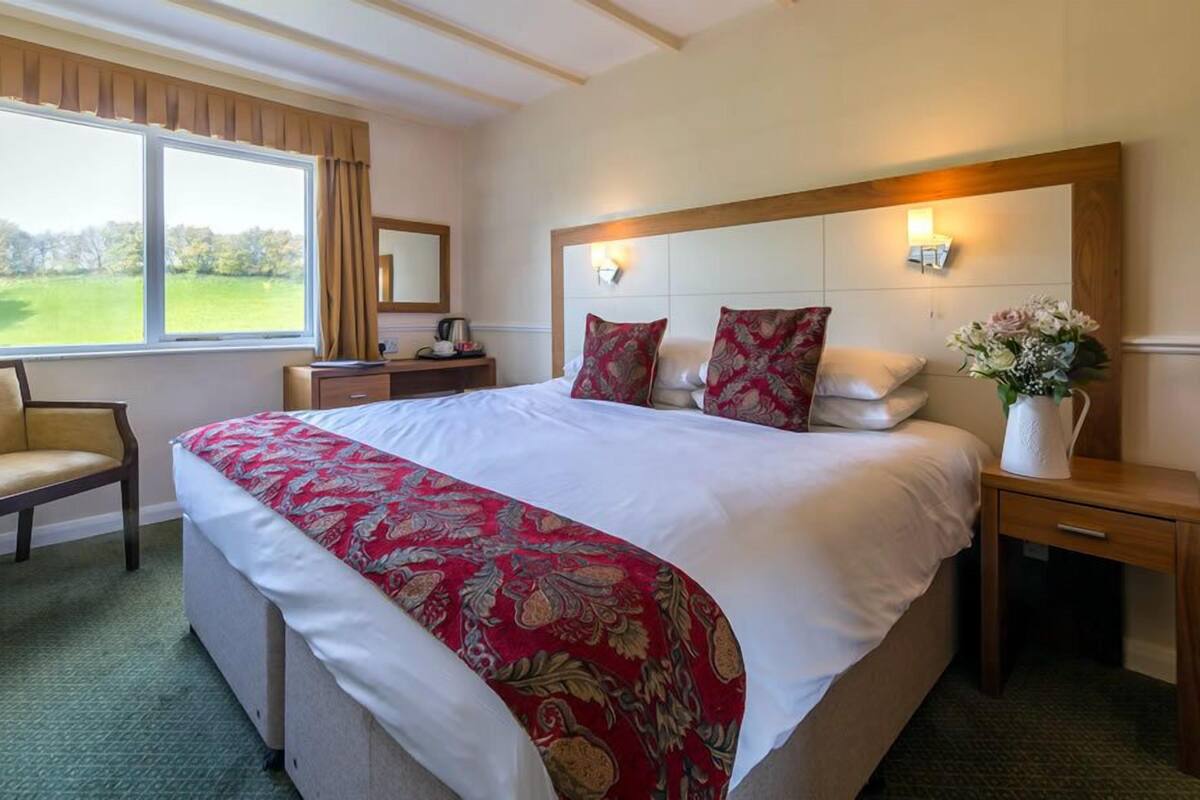 A hotel room with a large bed, a chair, a nightstand, a bedside table with a vase of flowers, and a window with curtains.