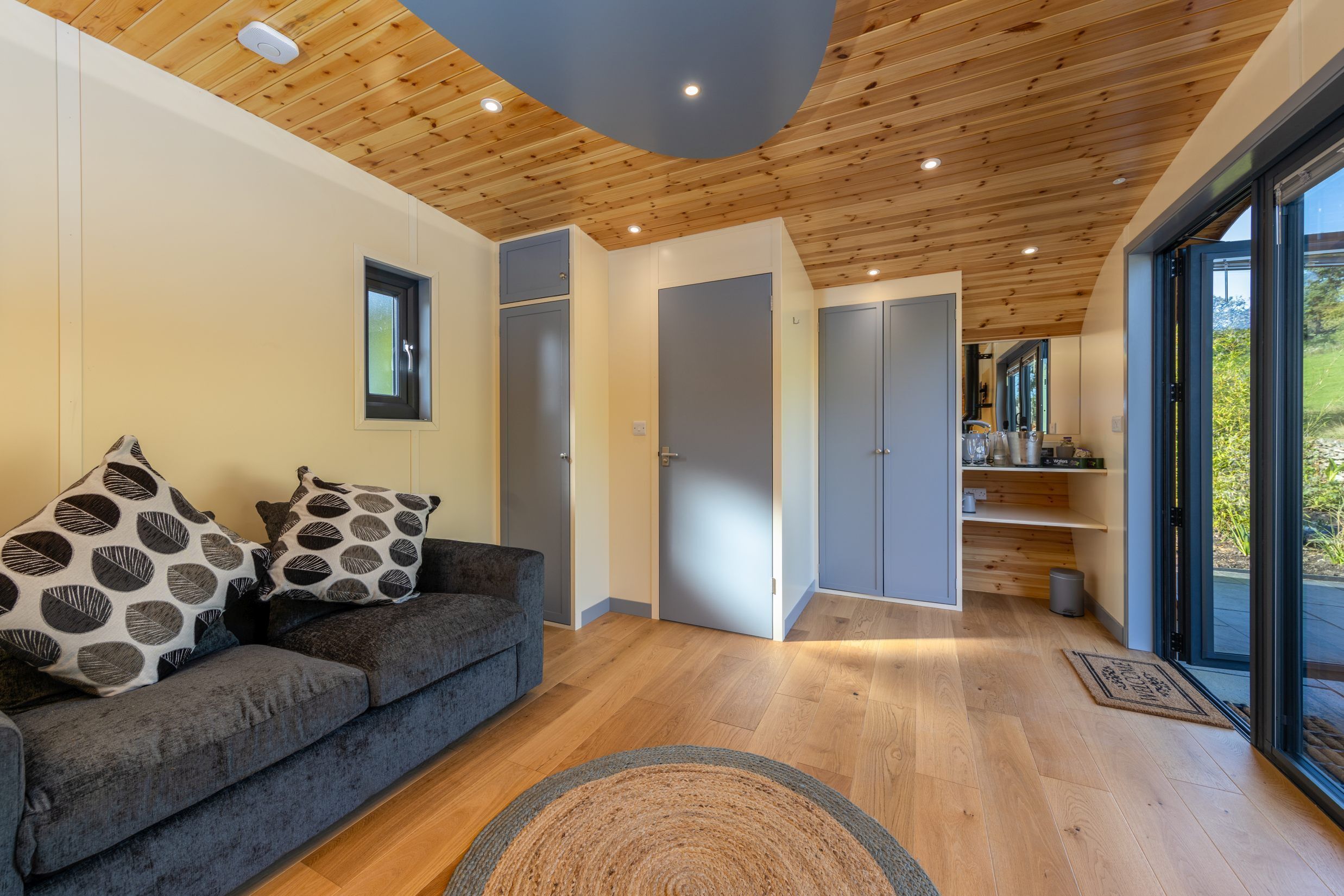 A modern interior with a wooden ceiling, grey sofa with patterned cushions, and a view through large glass doors.