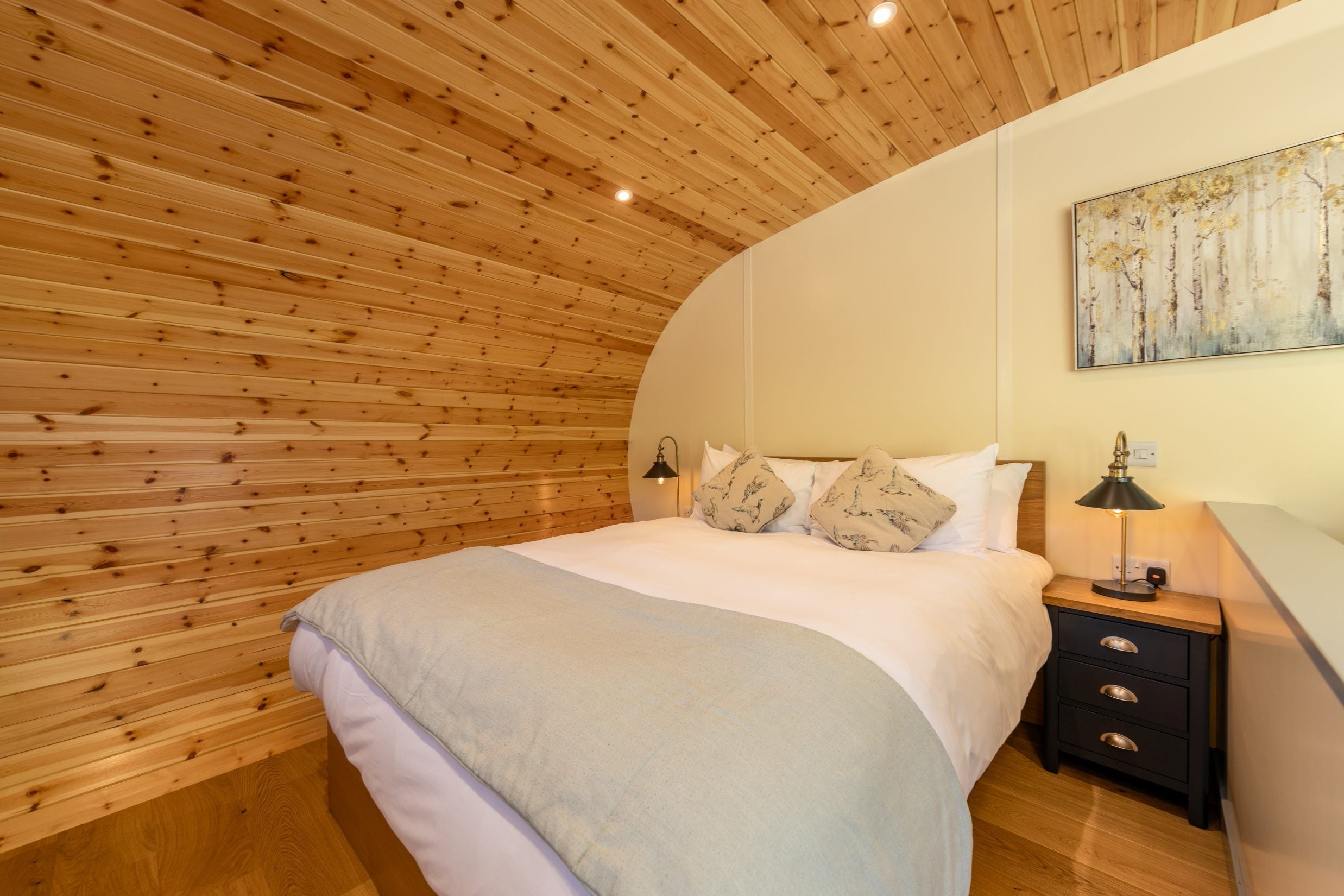 A bedroom with a wooden slat ceiling, a bed with white bedding, a nightstand with a lamp, and a painting on the wall.