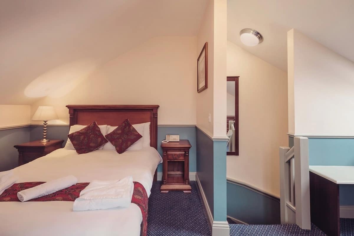 A hotel room with a large bed, a bedside table with a lamp, a small wooden nightstand, and a desk with a chair.