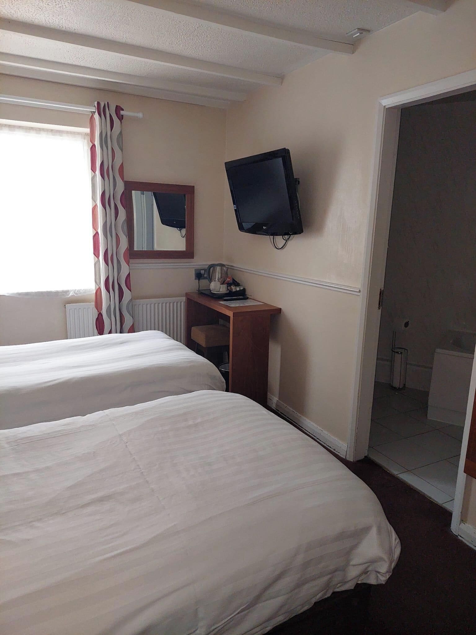 Habitación de hotel con dos camas, un televisor instalado en la pared, un escritorio pequeño con un hervidor y un espejo sobre el escritorio.