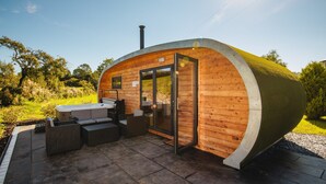 A modern, curved wooden cabin with a glass door, furnished with a sofa, chairs, and a hot tub. - Damson Dene Hotel (Kendal)