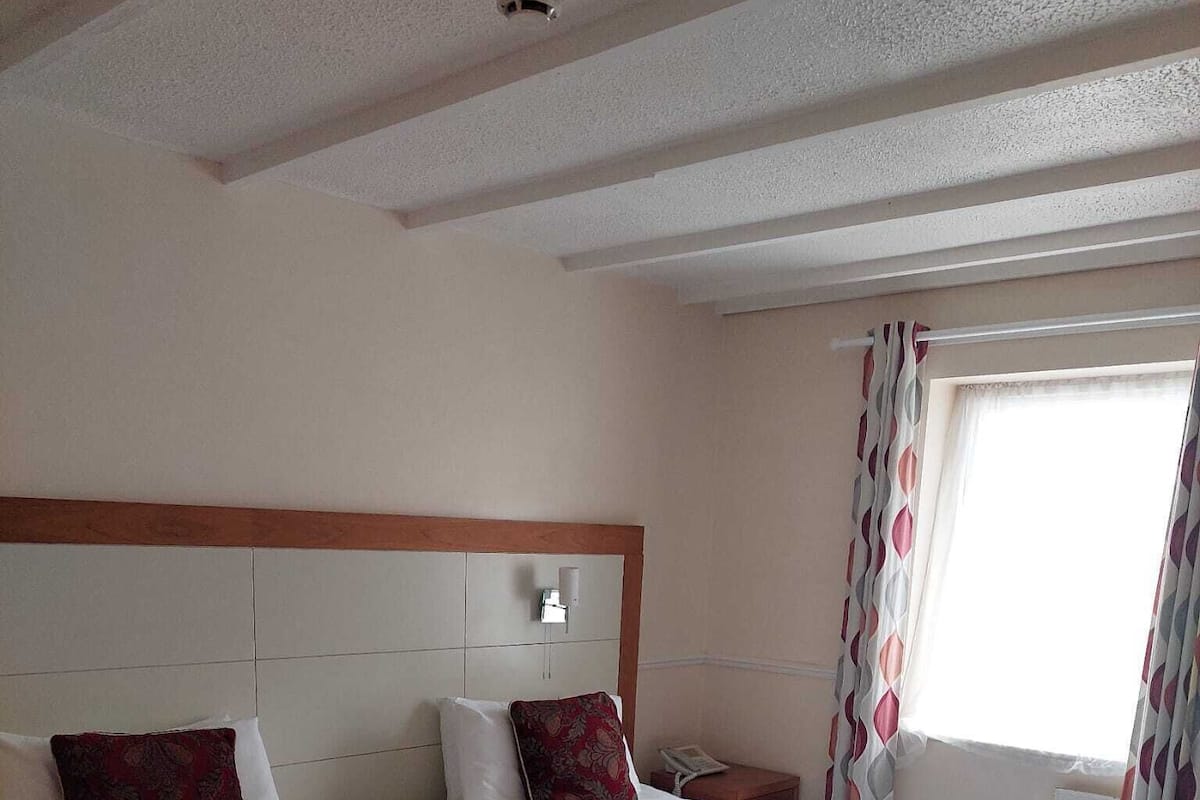 A hotel room with two beds, a headboard, a nightstand, a window with curtains, and a ceiling light.