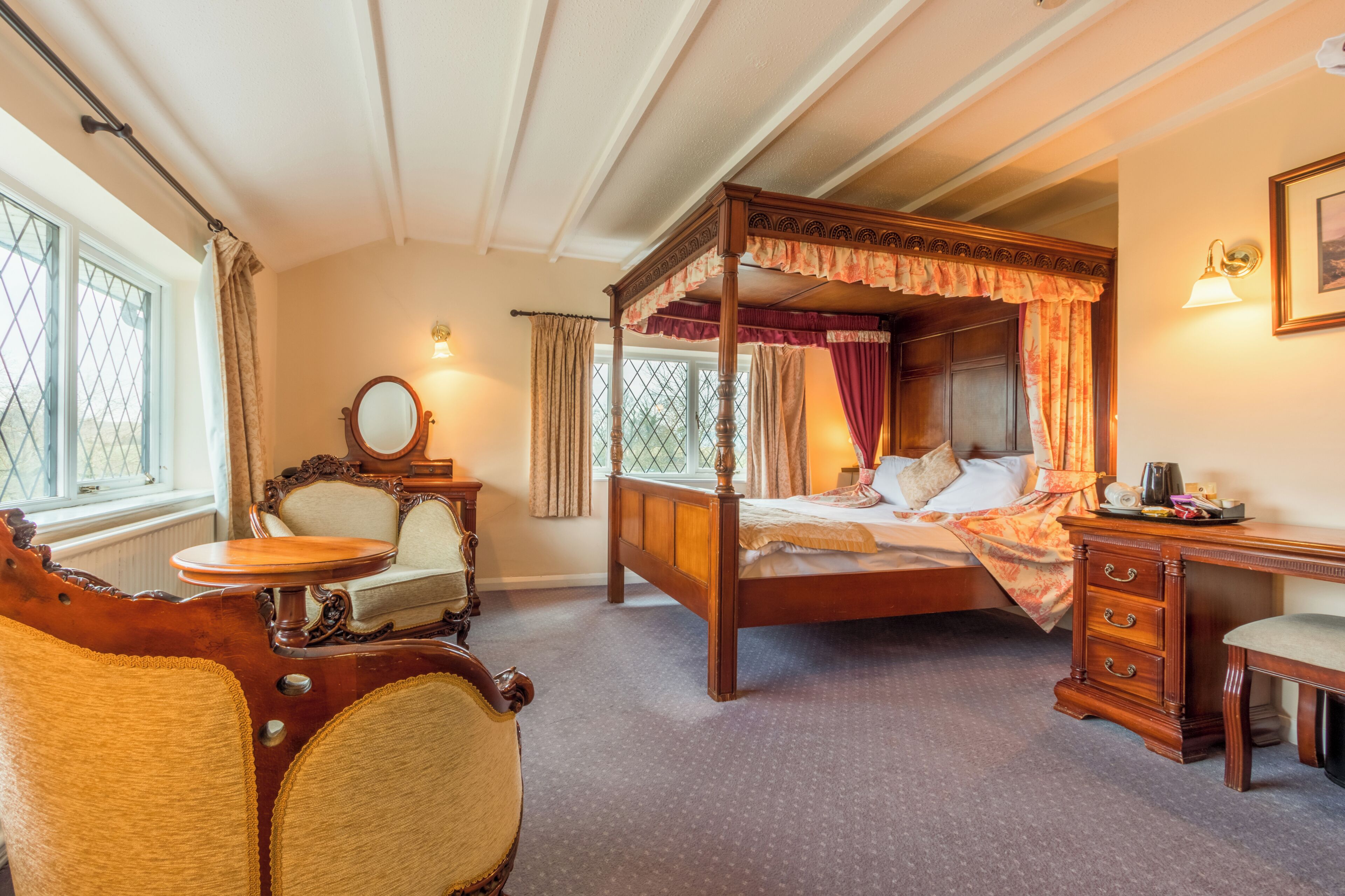 A four-poster bed, a wooden desk, a chair, and a small table in a room with a window and a radiator.