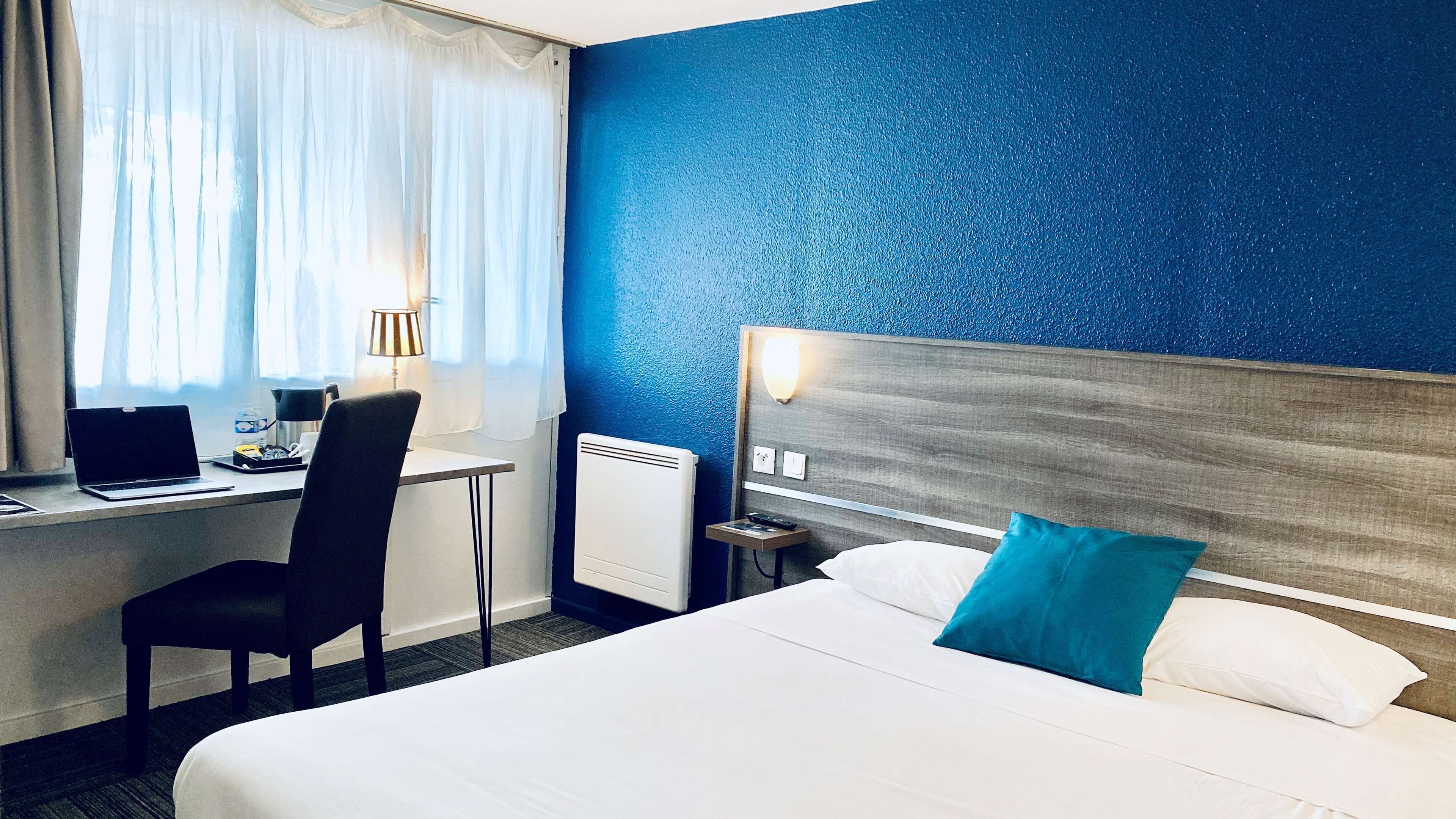 A hotel room with a bed, a desk with a laptop, a chair, a nightstand, and a blue textured wall.
