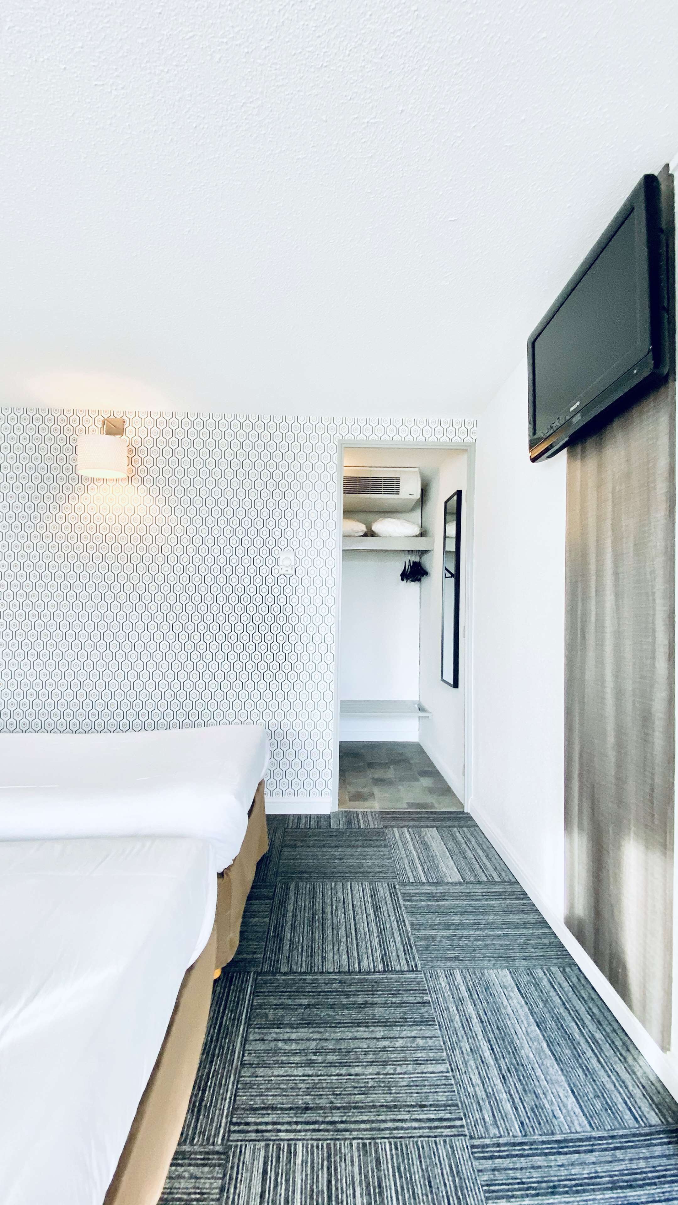 A hotel room with a bed, a TV mounted on the wall, a patterned wall, and a hallway leading to a bathroom.