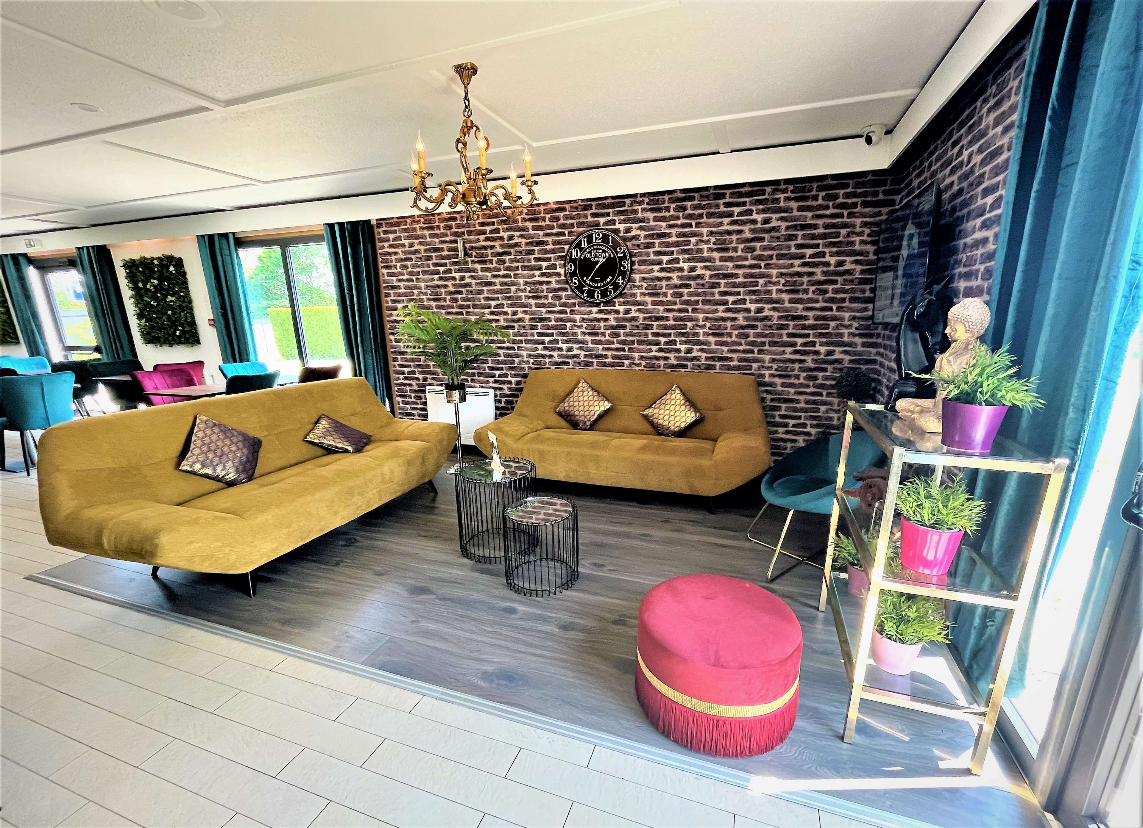 Un salon moderne avec deux canapés de couleur moutarde, un pouf rouge, un lustre noir et un mur en briques orné d’une horloge.