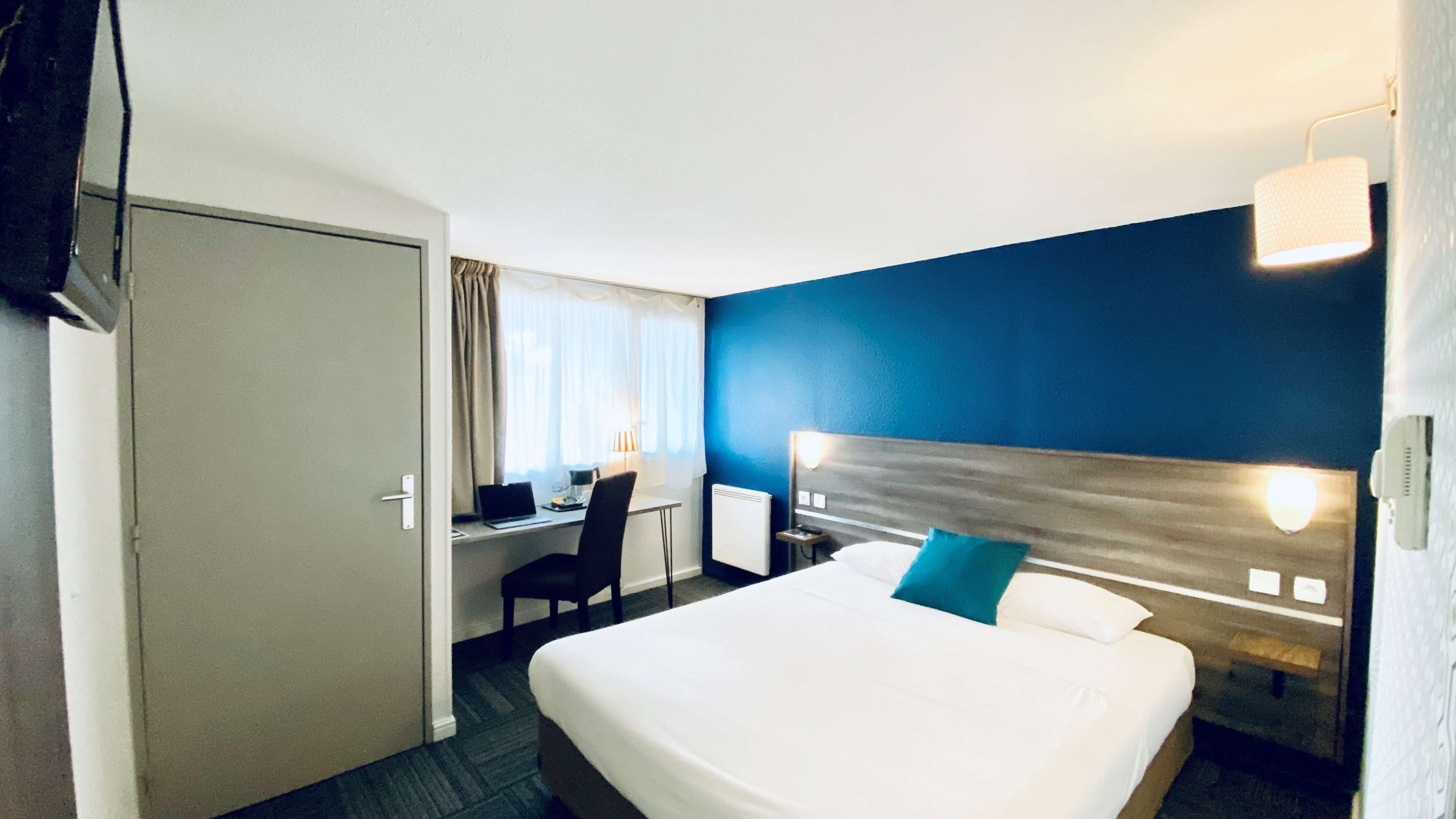 A hotel room with a bed, a desk with a laptop, a chair, a TV, and a nightstand with a lamp.