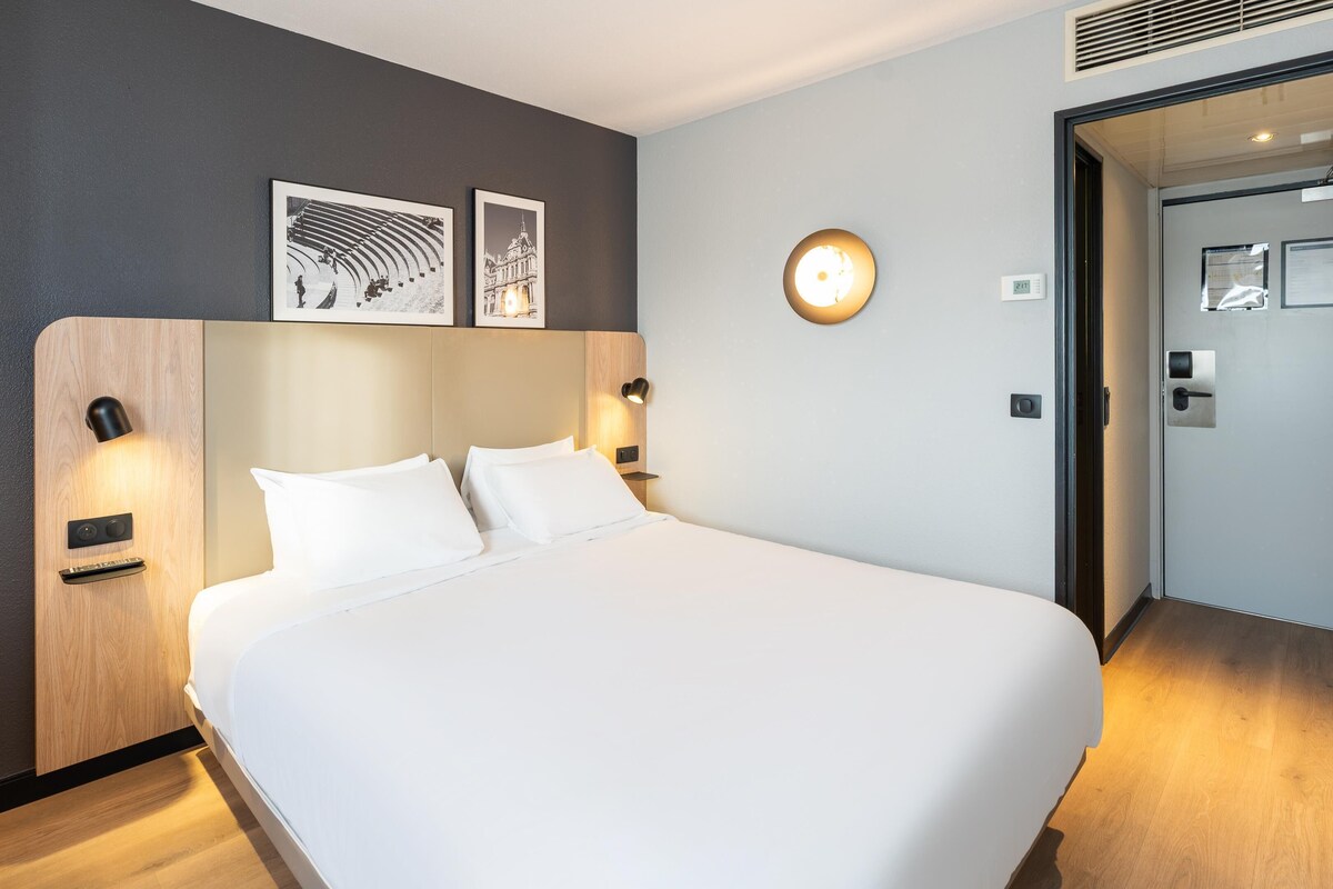 Une chambre d’hôtel avec un grand lit, un bureau et une chaise. Il y a deux lampes fixées au mur et une horloge accrochée au mur.