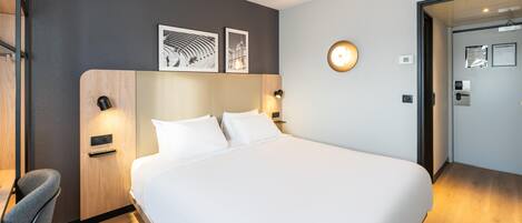 Habitación de hotel con una cama grande, un escritorio y una silla. Hay dos lámparas de pared y un reloj en la pared.