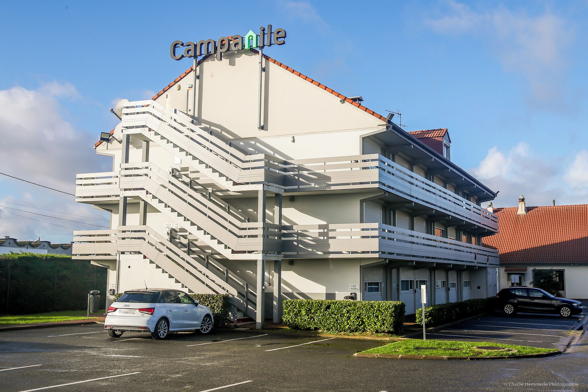 Un edificio de varios pisos con un letrero que dice "Carpanille". Tiene un estacionamiento con varios autos y una escalera que lleva a la entrada.
