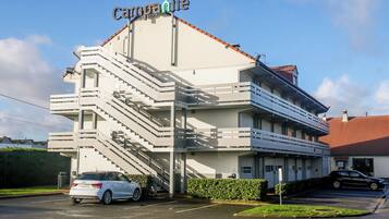 Un edificio de varios pisos con un letrero que dice "Carpanille". Tiene un estacionamiento con varios autos y una escalera que lleva a la entrada.