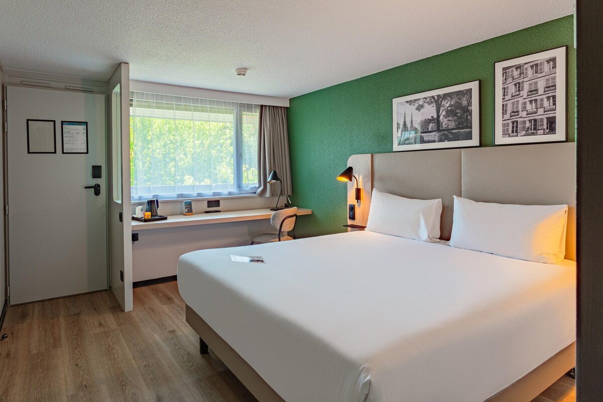 Une chambre d’hôtel comprenant un lit, un bureau avec une chaise, une fenêtre avec des rideaux et deux tableaux encadrés accrochés au mur.