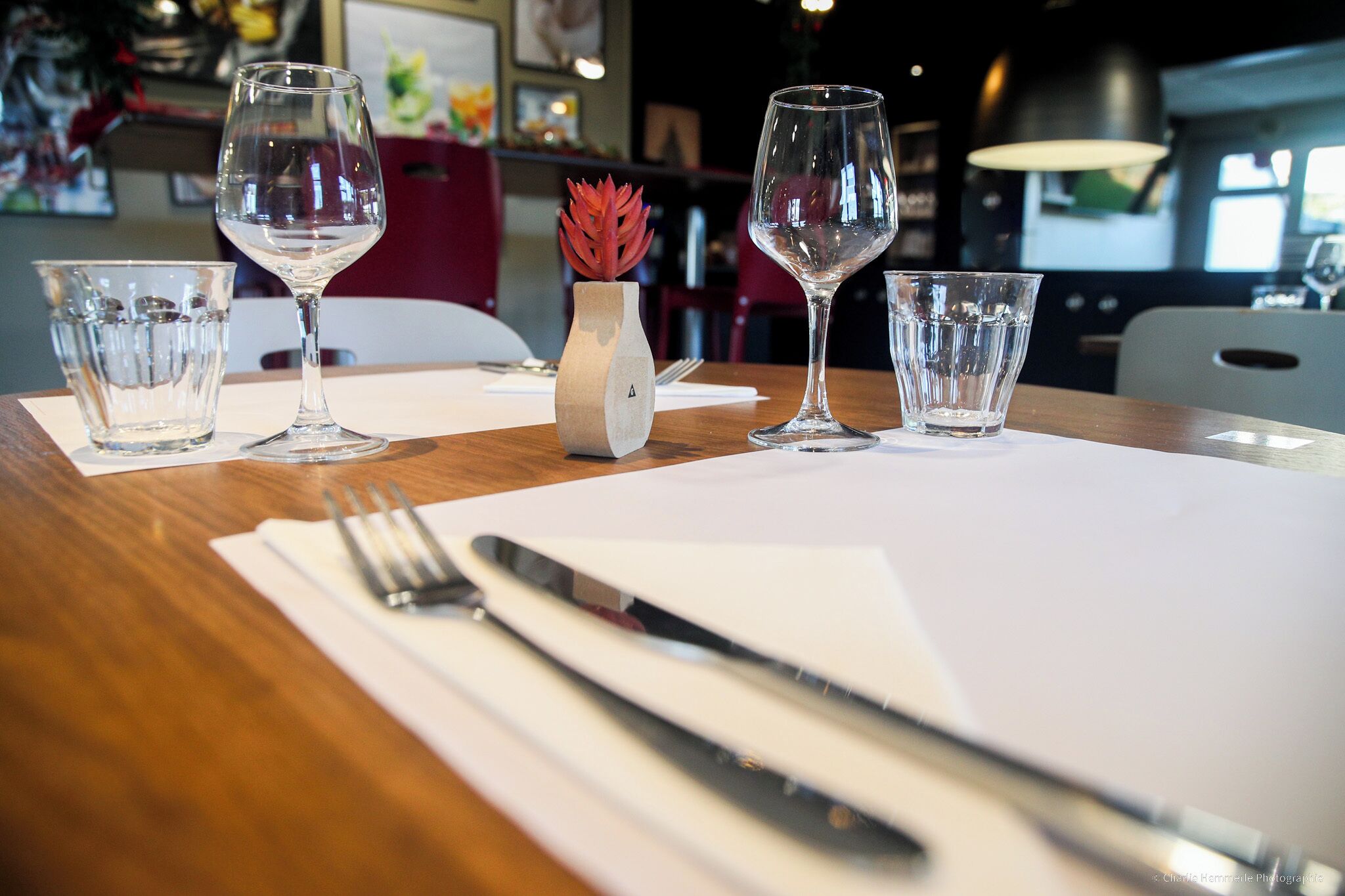Una mesa de comedor con copas de vino, un florero con una flor roja, y un cuchillo y tenedor sobre un servilleta blanca.