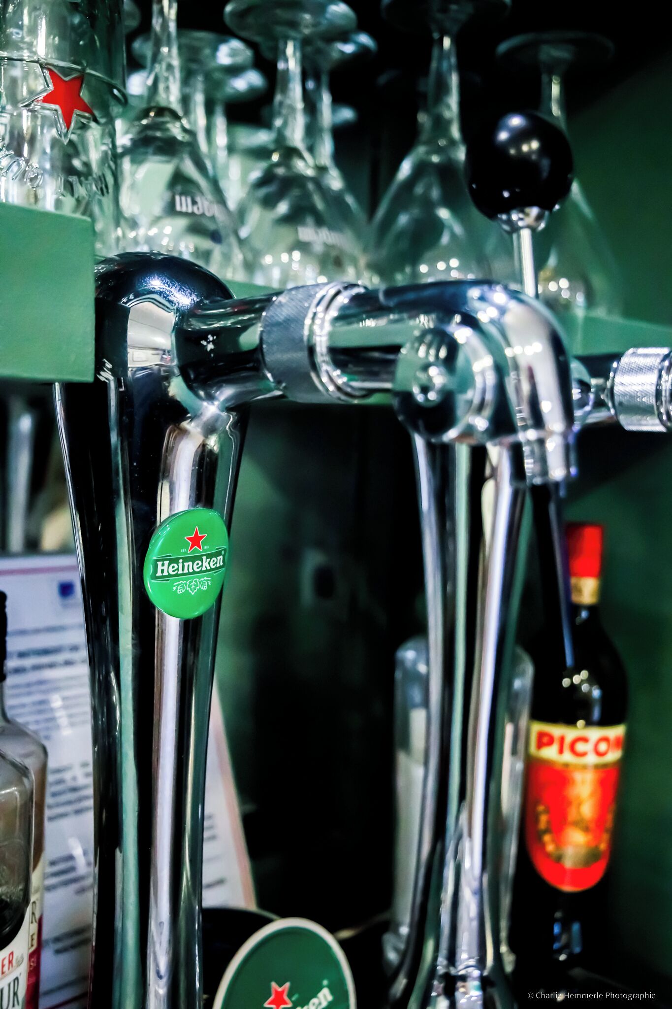 Uma torneira de bar com rótulo Heineken, uma garrafa de Picon e vários copos.