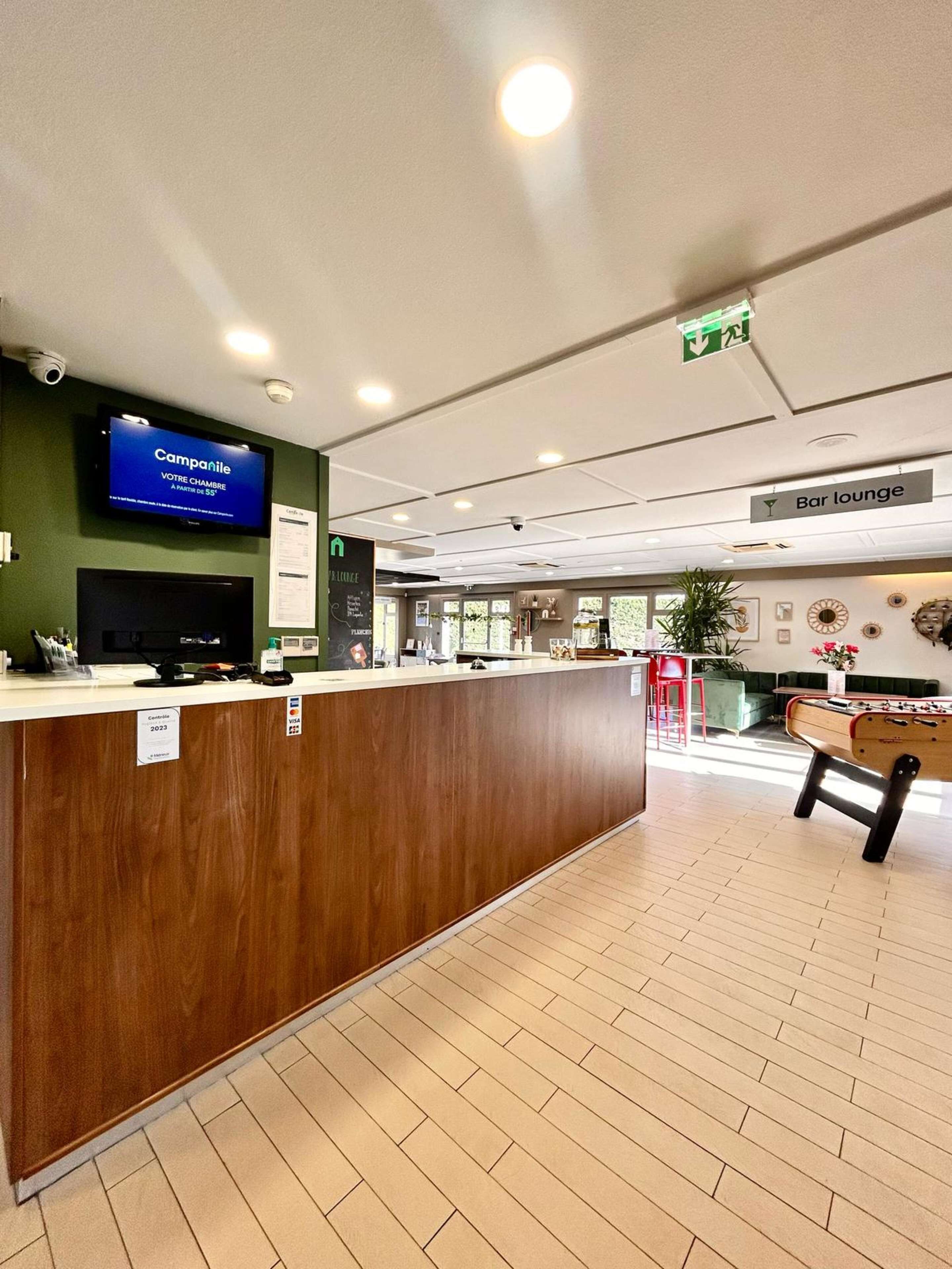 Un espace d’accueil avec un comptoir en bois, un téléviseur diffusant des images de « Campanile » et une enseigne indiquant « Bar lounge ».