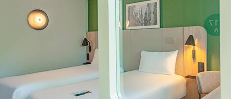 A hotel room with two beds, a wall-mounted lamp, a bedside table, and a framed picture on the wall.
