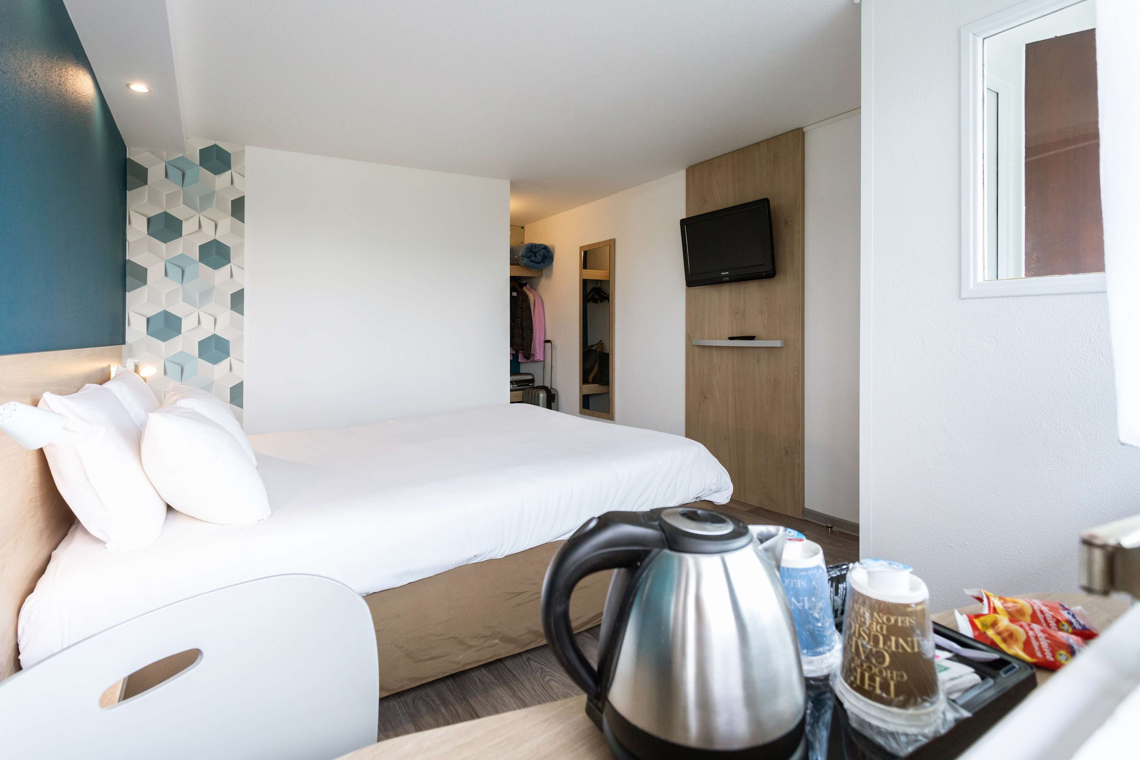A hotel room with a bed, a kettle, a tray with snacks, a TV, and a geometric wall design.