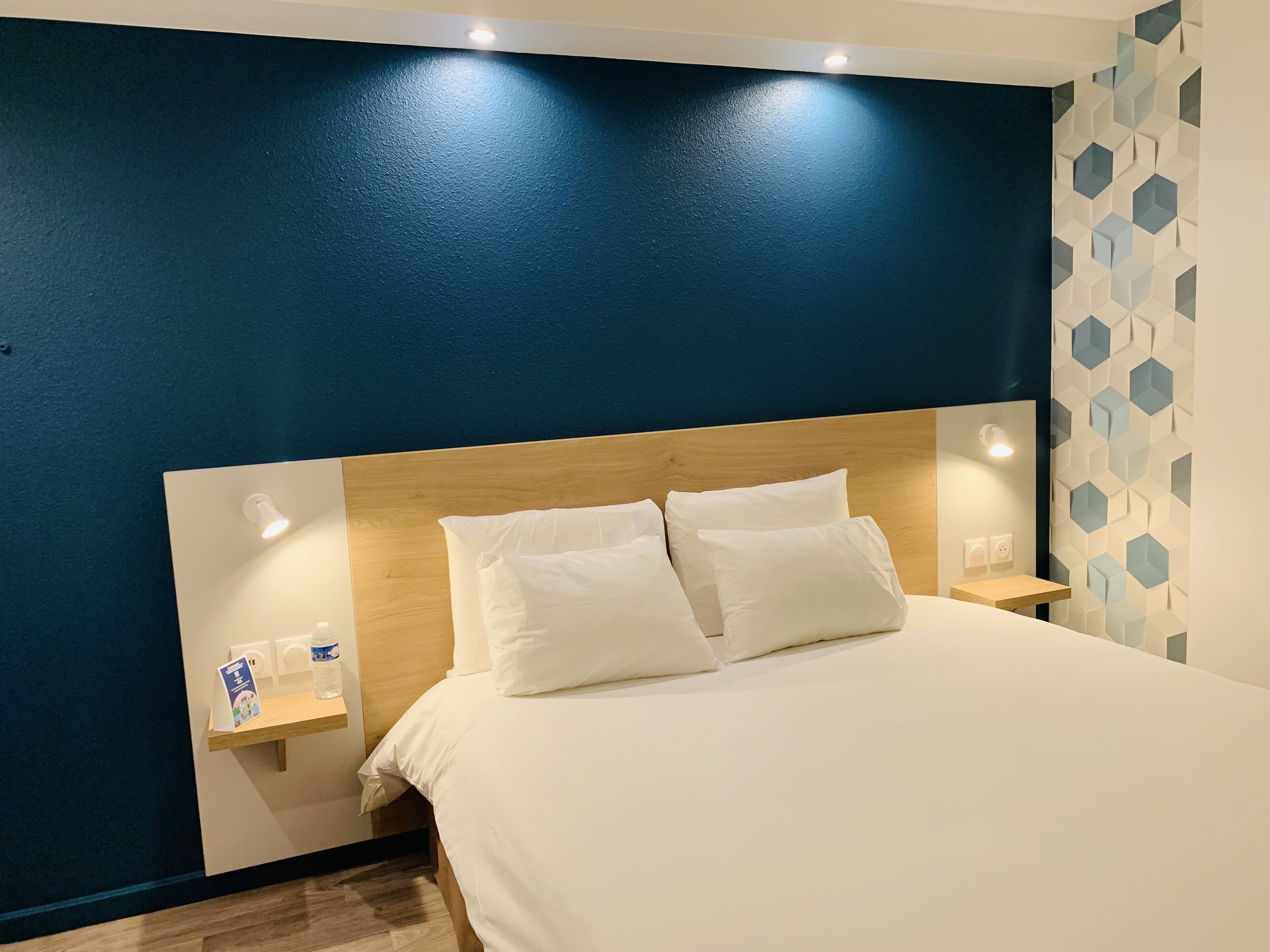 Een hotelkamer met een groot bed, een houten hoofdbord, nachtkastjes met lampen en een wand met een geometrisch patroon.