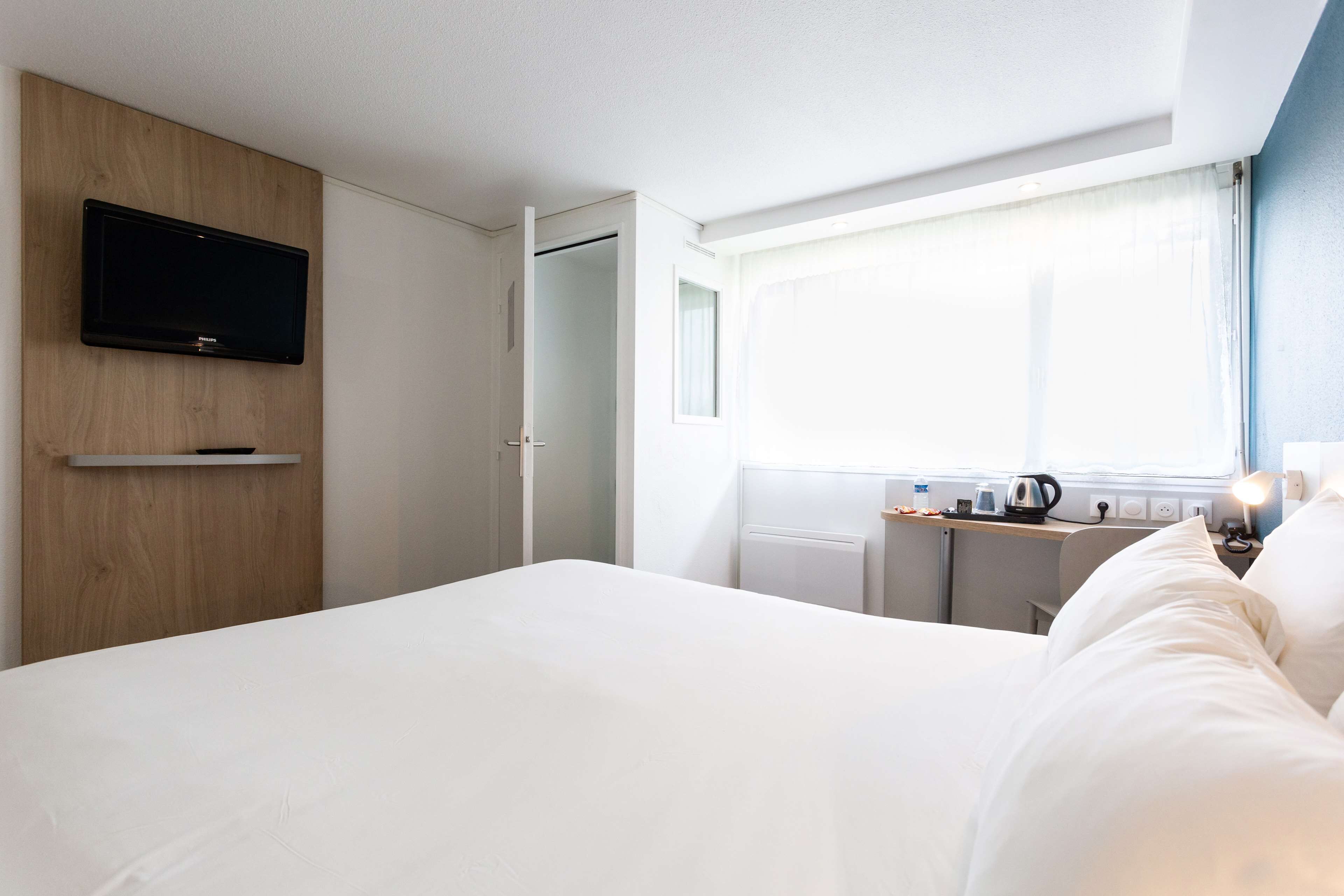 A hotel room with a bed, a TV mounted on a wooden panel, a desk with a kettle, and a window with curtains.