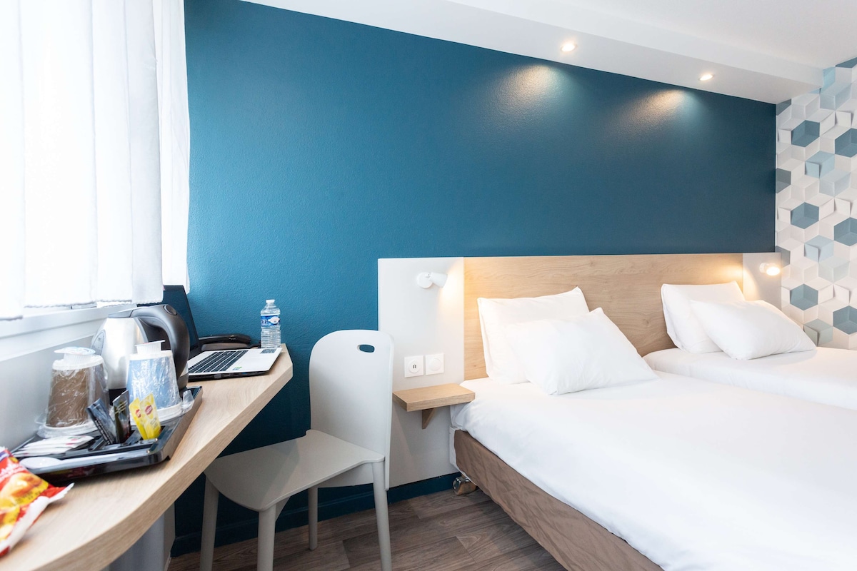 Habitación de hotel con cama, escritorio, laptop, silla y ventana con cortinas.