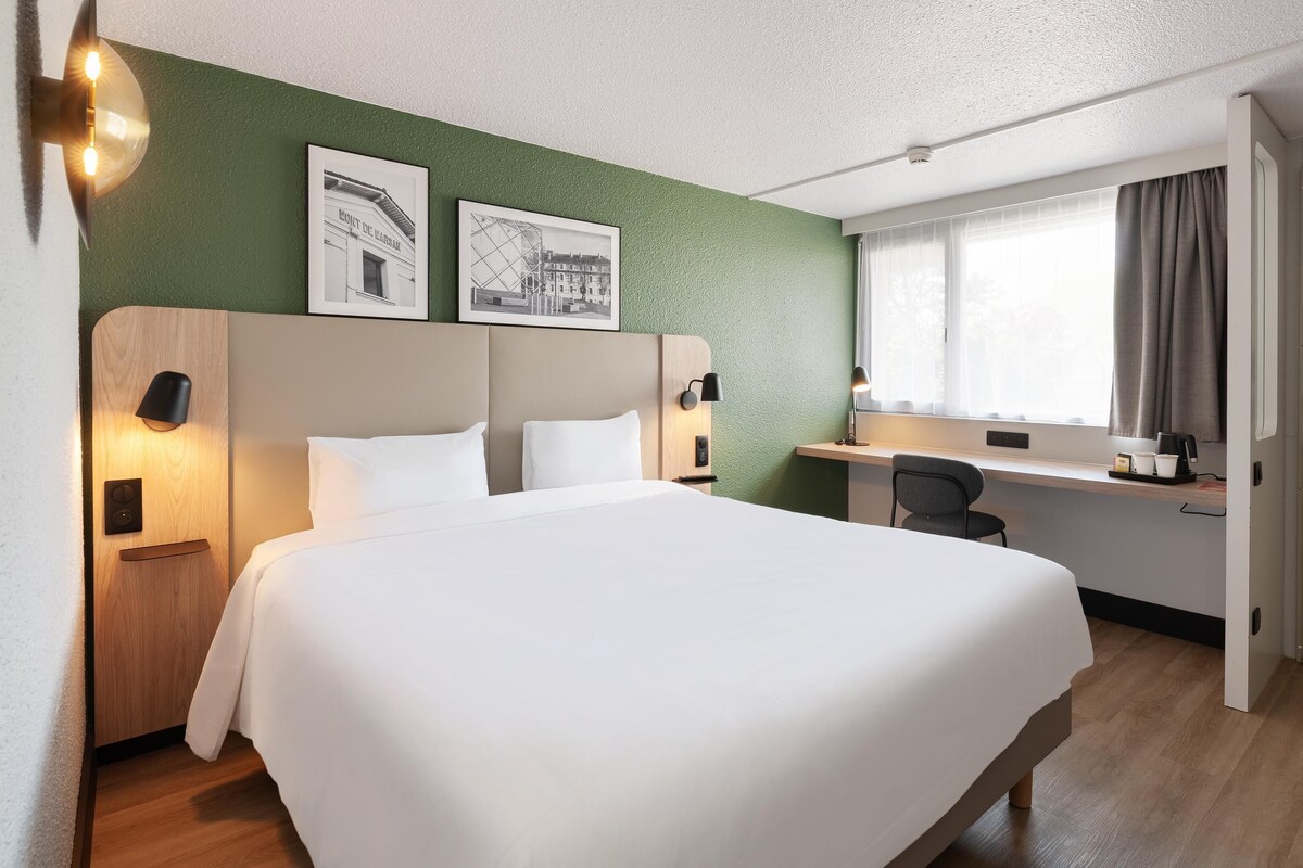 A hotel room with a large bed, a desk, a chair, and a window with curtains.