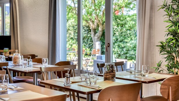 A restaurant with tables set for dining, large windows offering a view of greenery, and a plant on the right side. - Campanile NATURE - Epône (Epone)