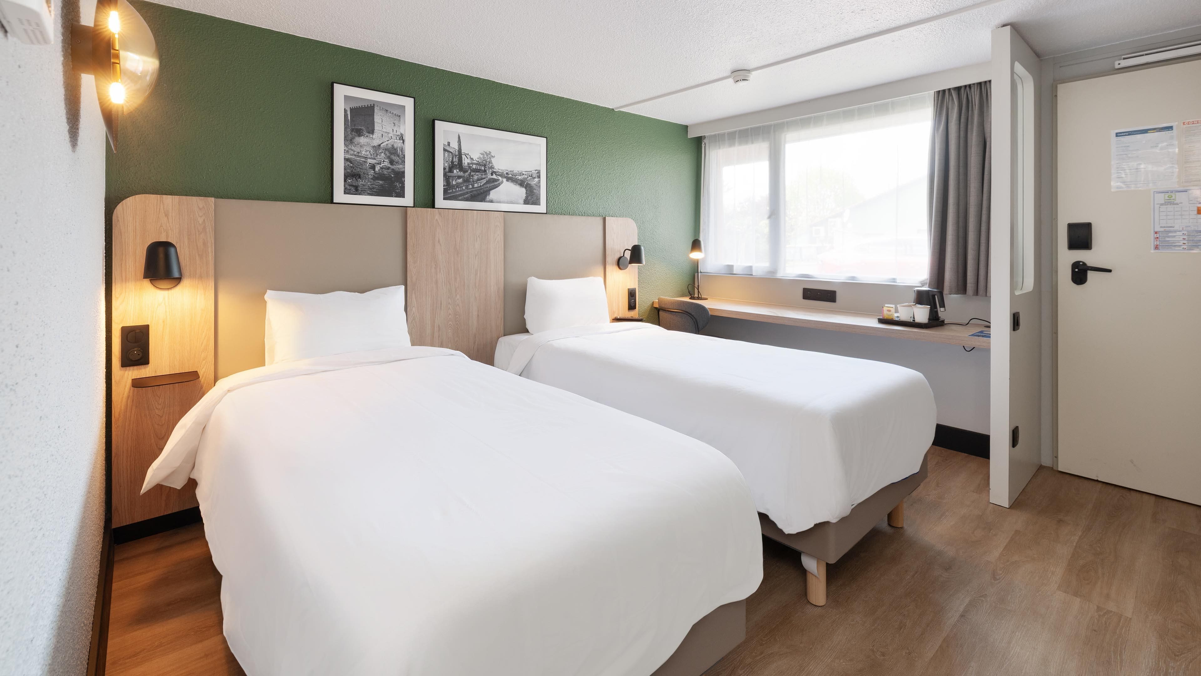 Une chambre d’hôtel avec deux lits, un bureau, une fenêtre avec des rideaux et deux tableaux encadrés au mur.