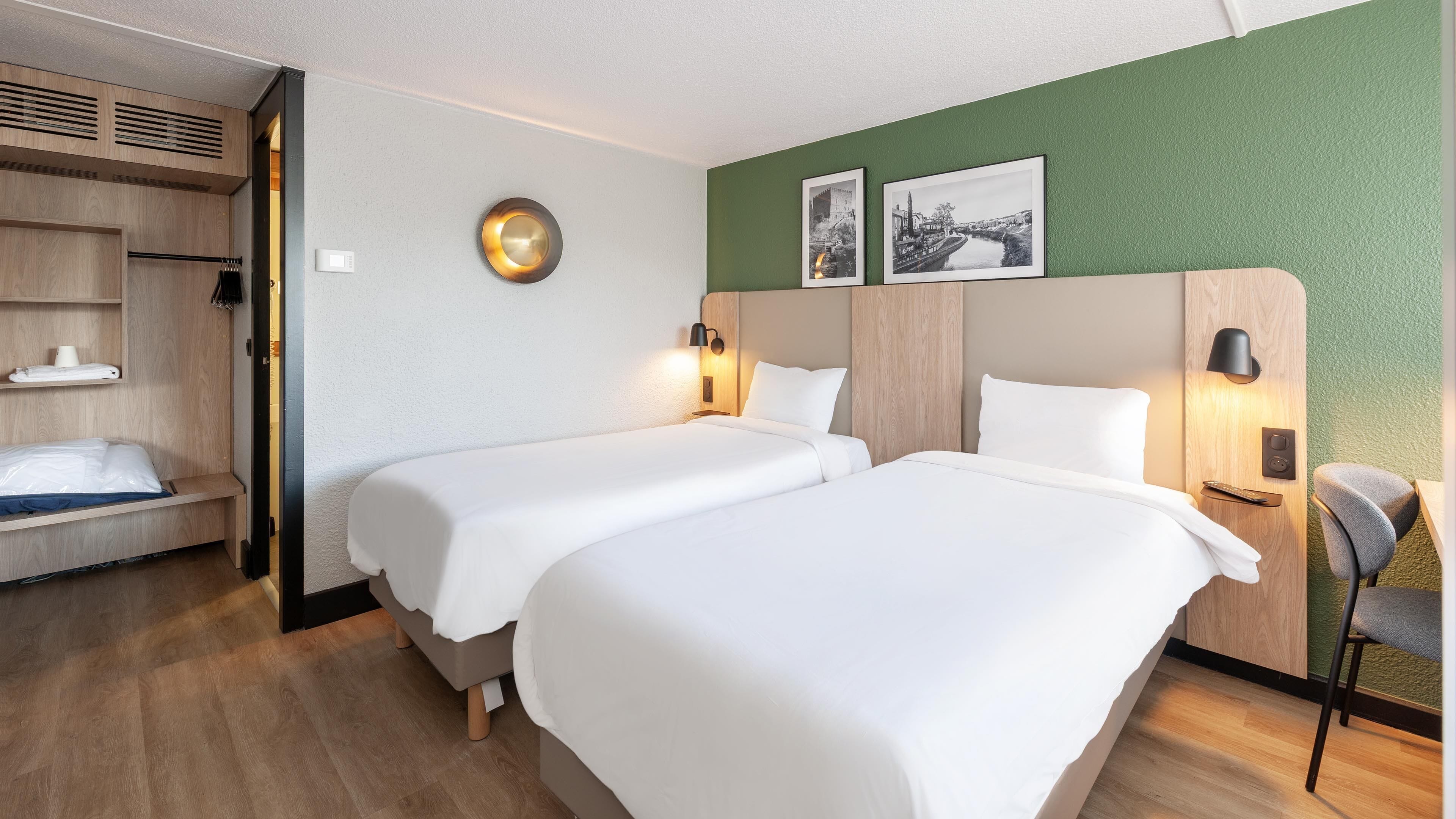 A hotel room with two beds, a wooden wardrobe, a chair, and a green wall with two framed pictures.