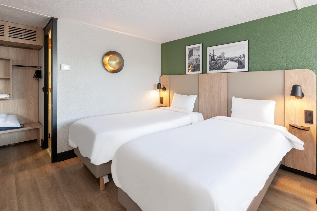 A hotel room with two beds, a wooden wardrobe, a chair, and a green wall with two framed pictures.