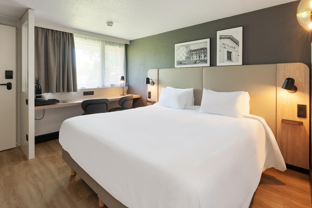 A hotel room with a large bed, a desk with a chair, a computer, and a window with curtains.