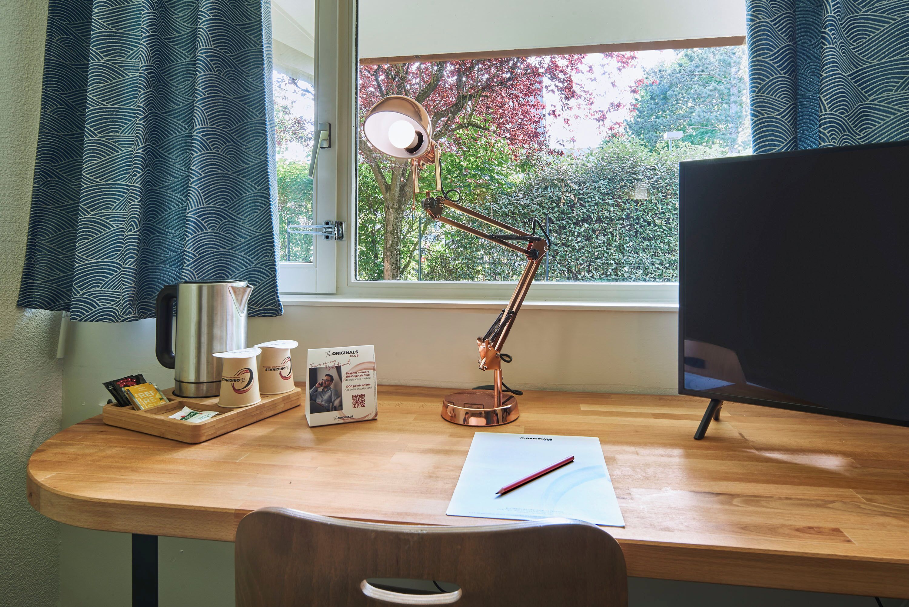 A wooden desk with a lamp, a computer monitor, a cup holder, and a window with curtains.