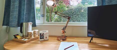 A wooden desk with a lamp, a computer monitor, a cup holder, and a window with curtains.