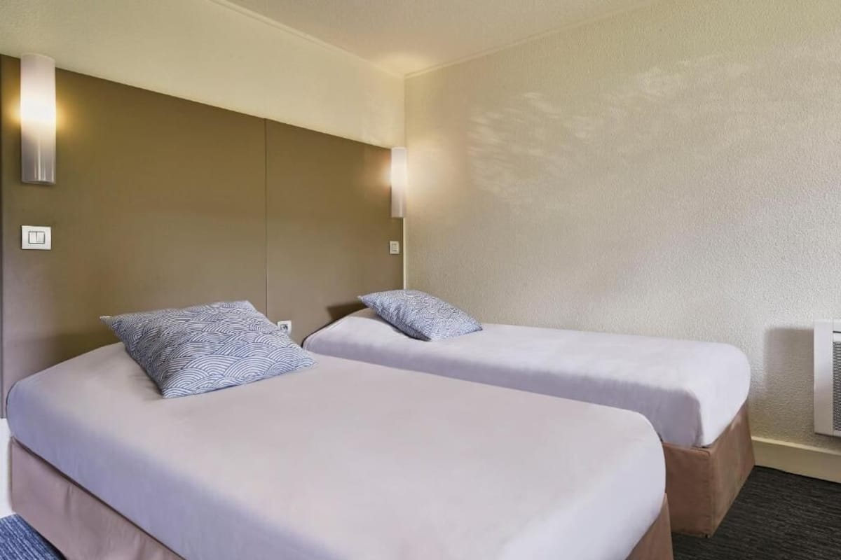 Ein Hotelzimmer mit zwei Einzelbetten, jedes mit einem gemusterten Kopfkissen, einer Wandleuchte und einem an der Wand angebrachten Thermostat.