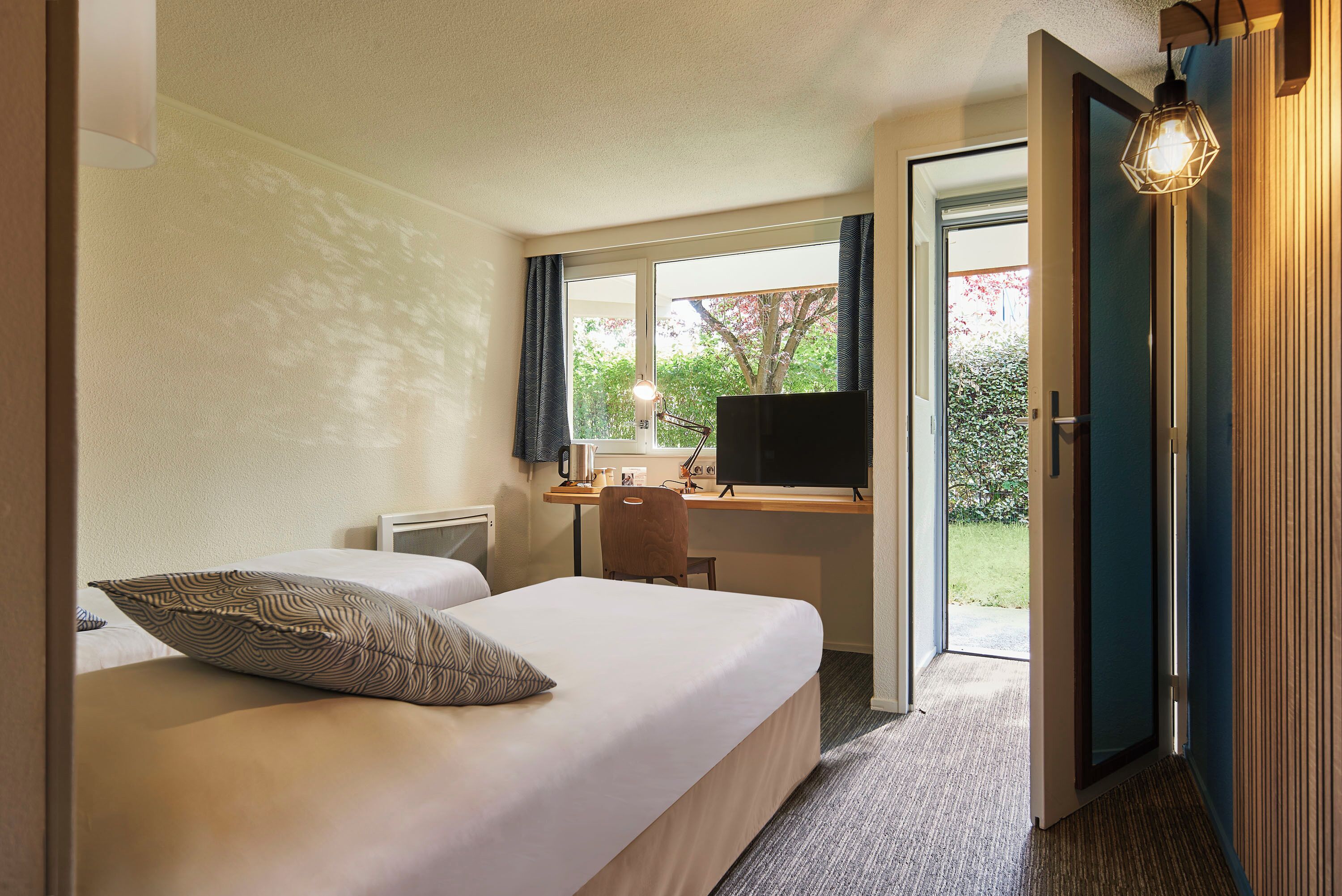 A hotel room with a bed, a desk with a computer, a chair, and a TV. There is a window with curtains and a view of greenery outside.