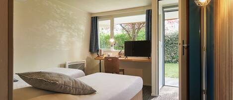 A hotel room with a bed, a desk with a computer, a chair, and a TV. There is a window with curtains and a view of greenery outside.