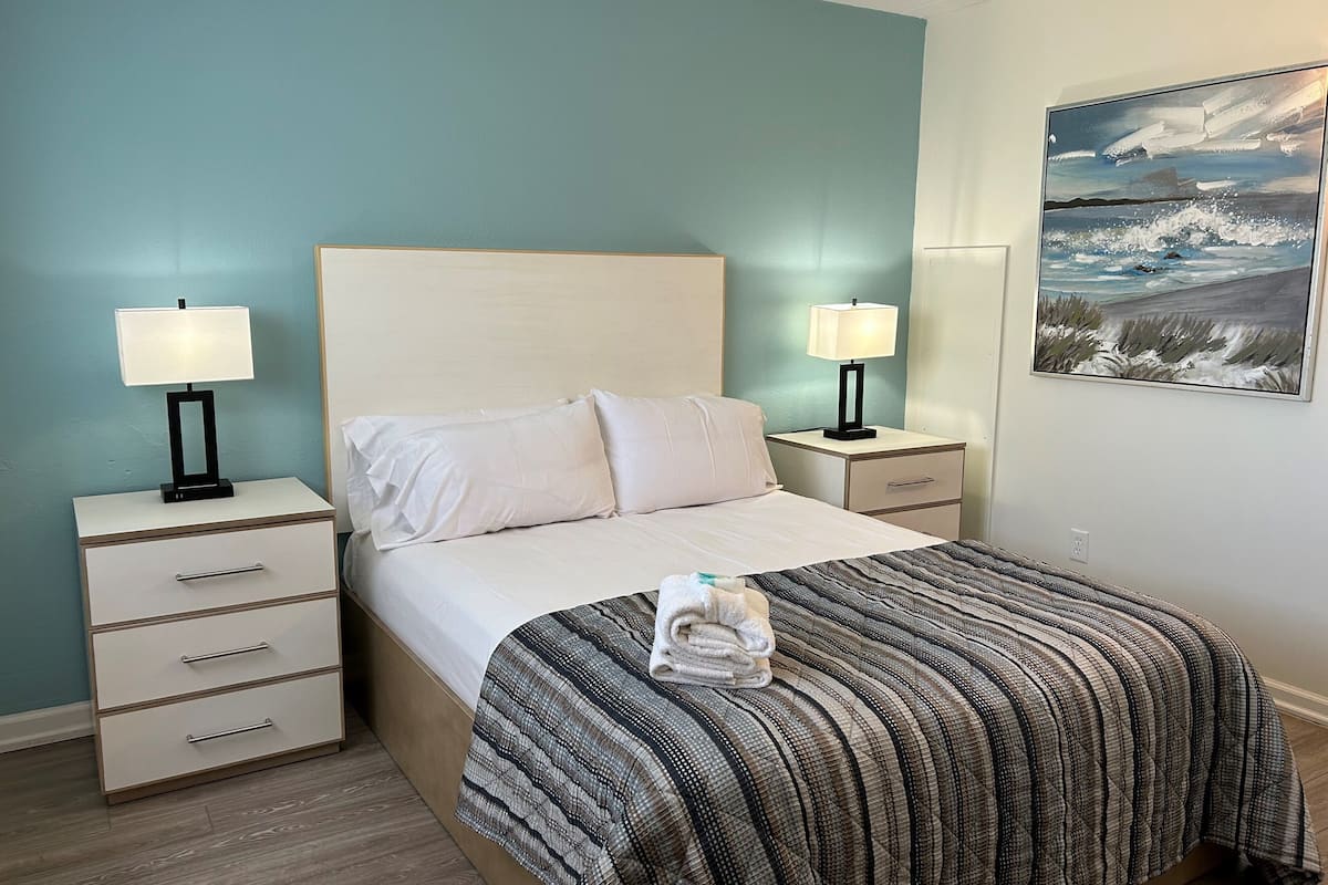 A neatly made bed with a striped blanket, two bedside tables with lamps, a nightstand with a drawer, and a painting on the wall.