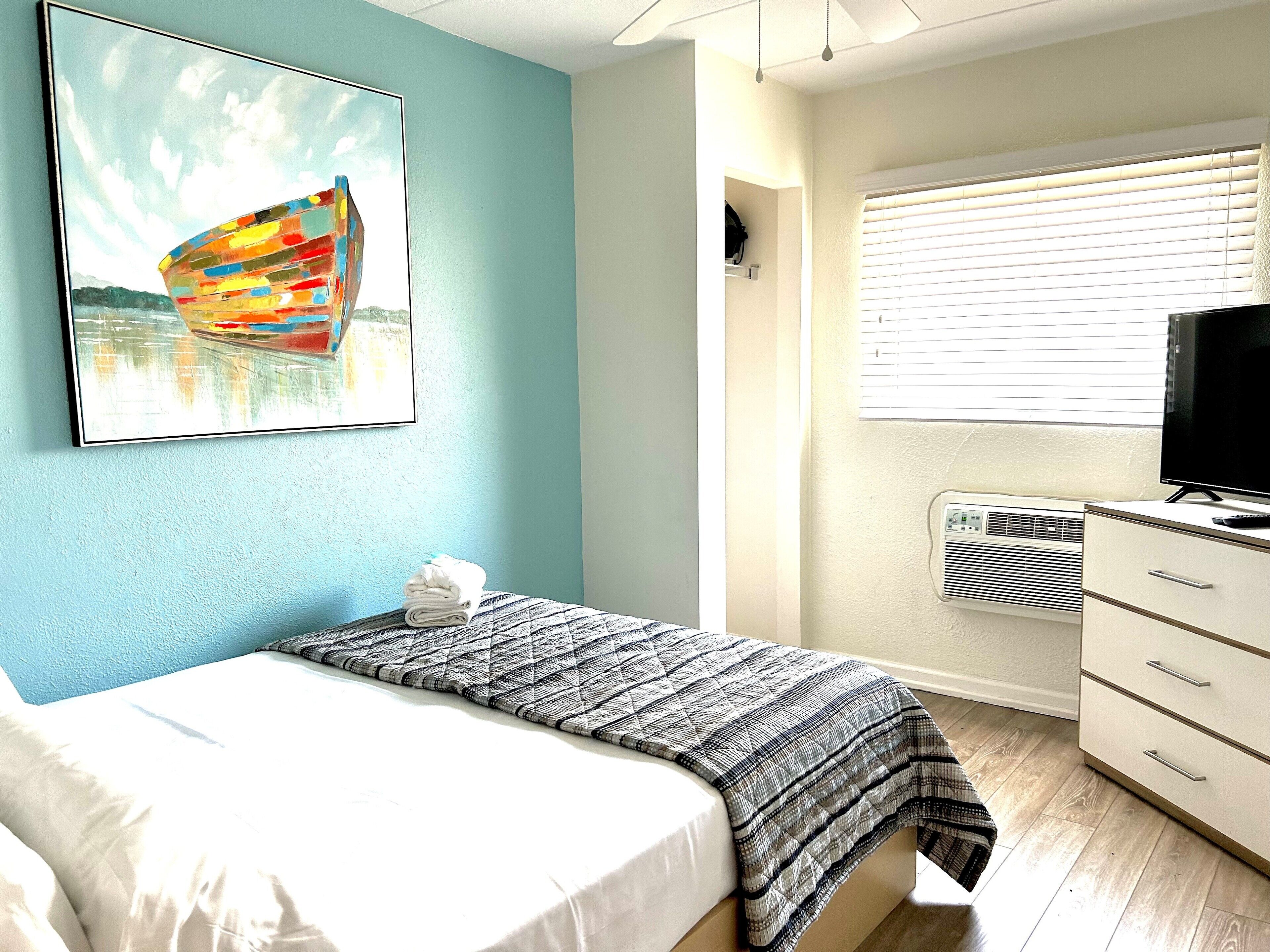 A bedroom with a bed, a window with blinds, a TV, and a painting on the wall.