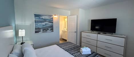 Et hotelværelse med seng, fjernsyn, kommode, et billede og et badeværelse, der kan ses gennem en åben dør.
